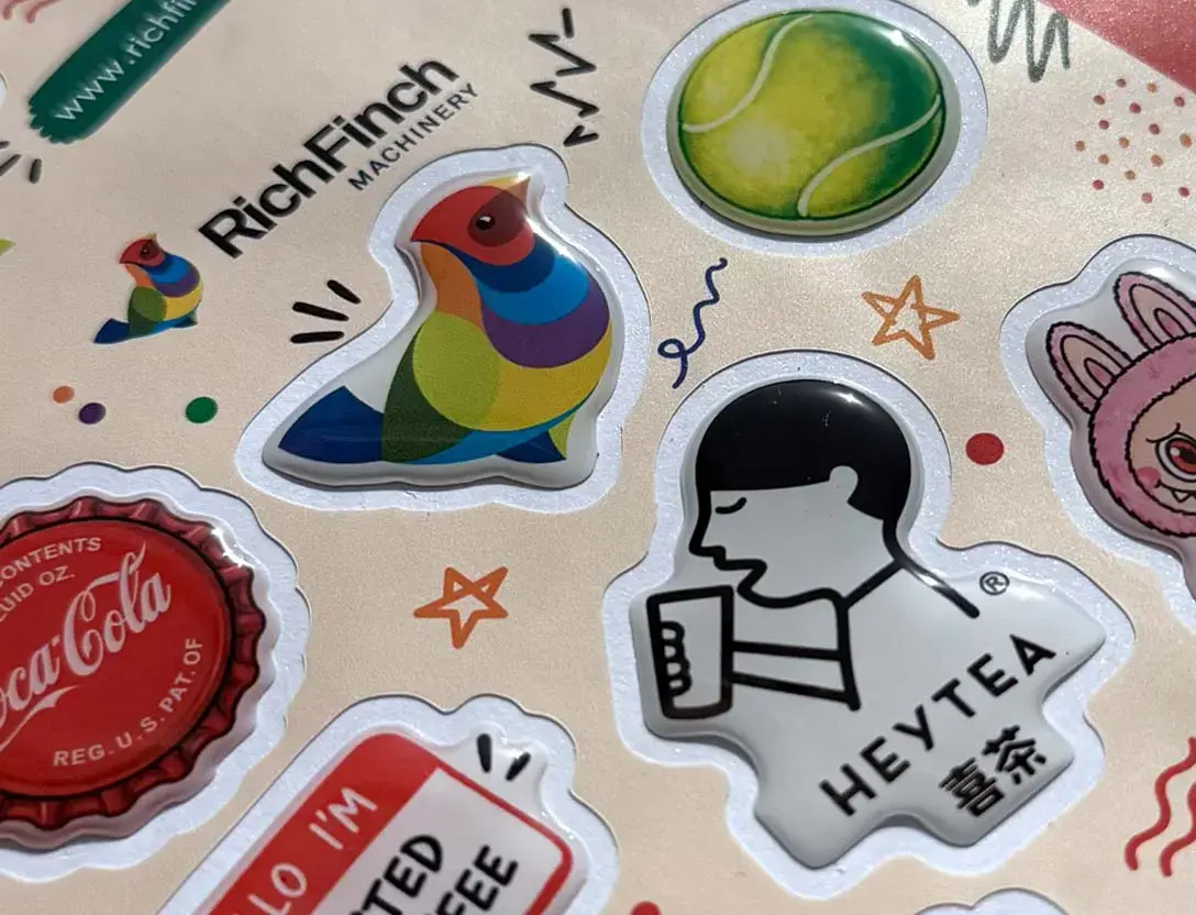 Clear resin domes on a sheet of printed stickers in cross-light, gloss highlights visible