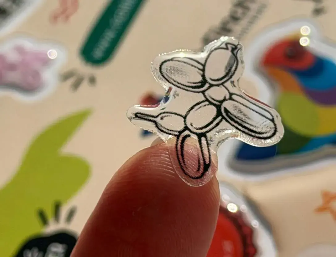 A single clear domed sticker balanced on a fingertip, a sheet of domed stickers blurred behind it